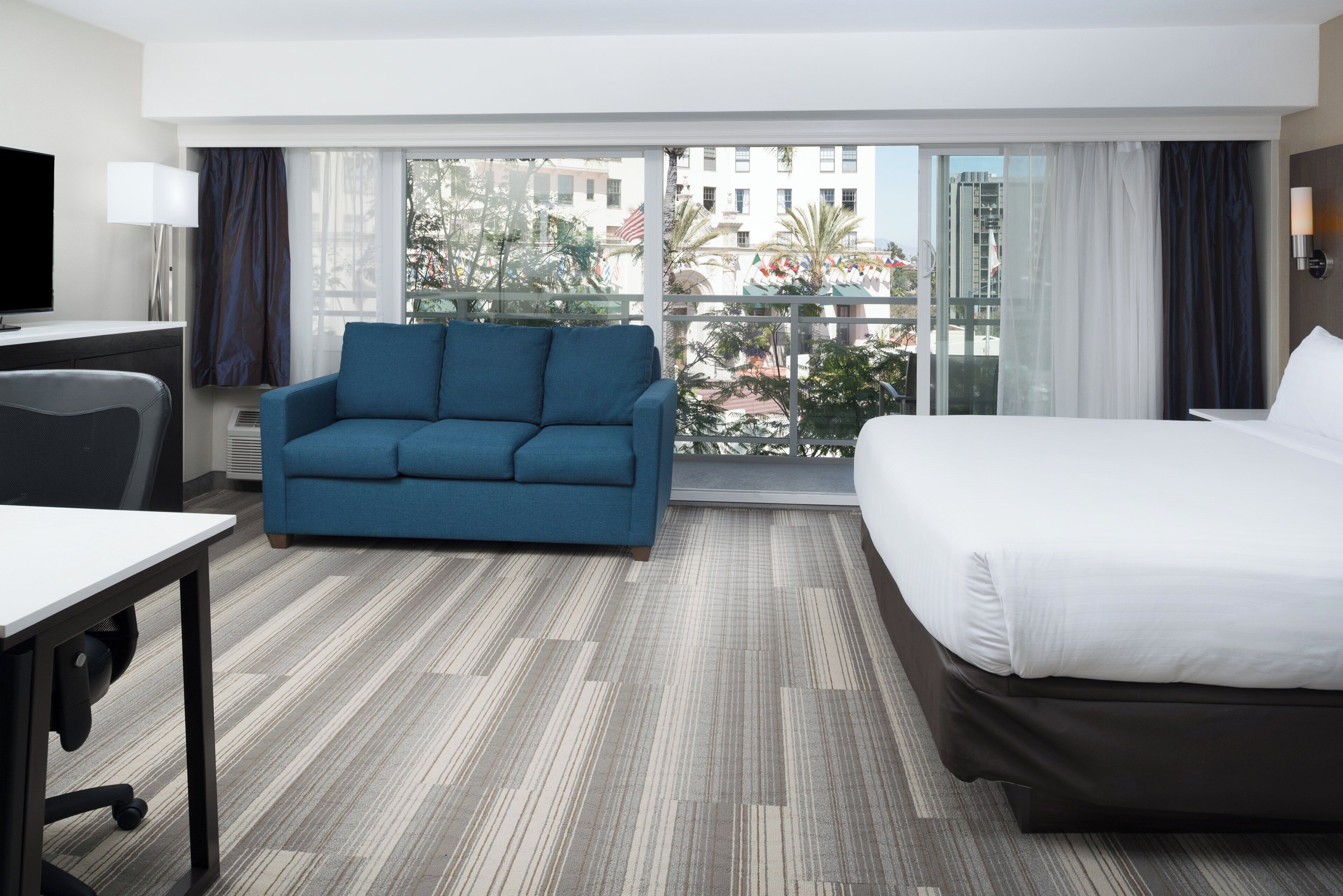 Une chambre d’hôtel comprenant un lit, un canap é, un bureau et une grande fenêtre donnant sur des immeubles et des arbres.