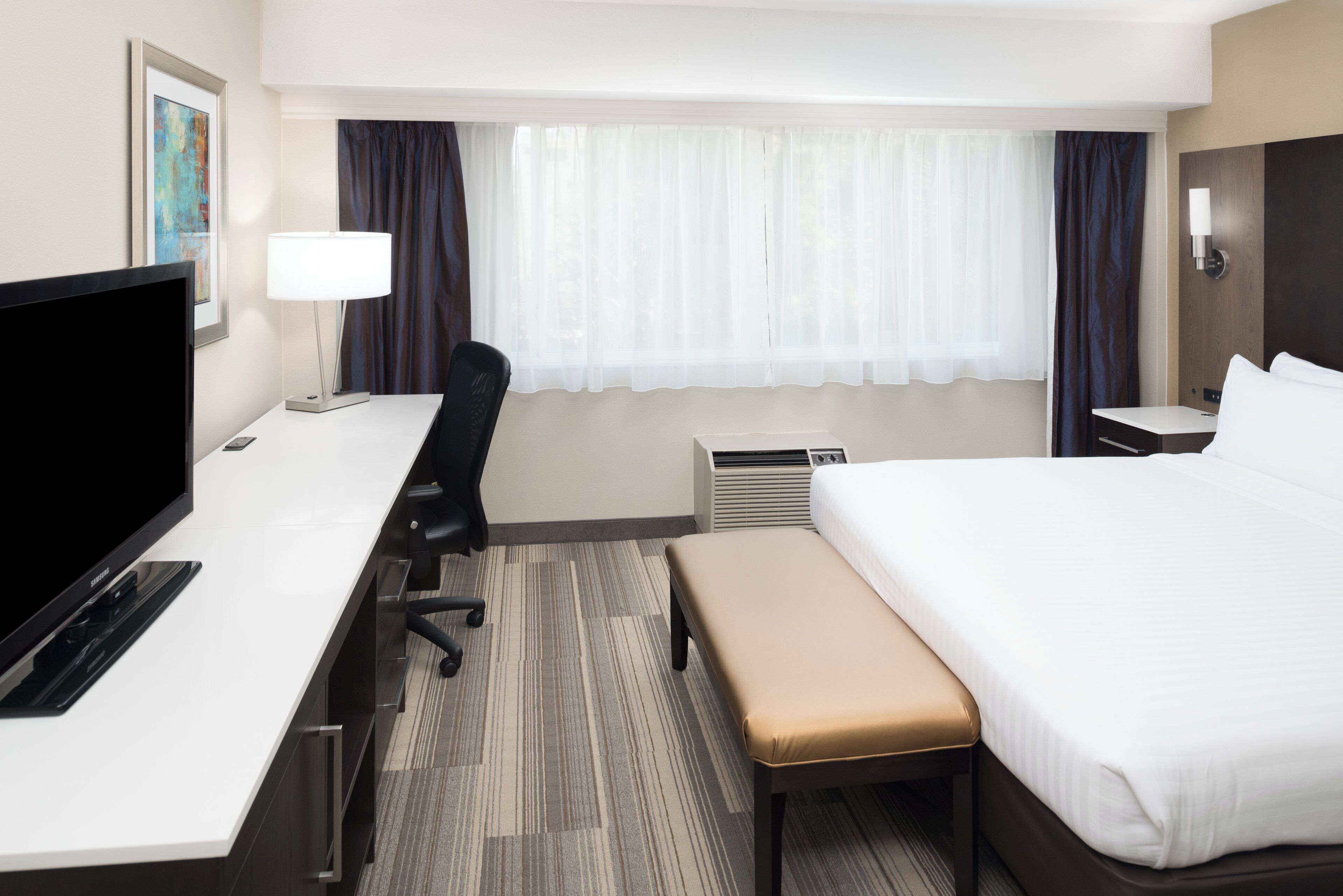 Habitación de hotel con cama, escritorio, silla, televisor, lámpara y ventana con cortinas.