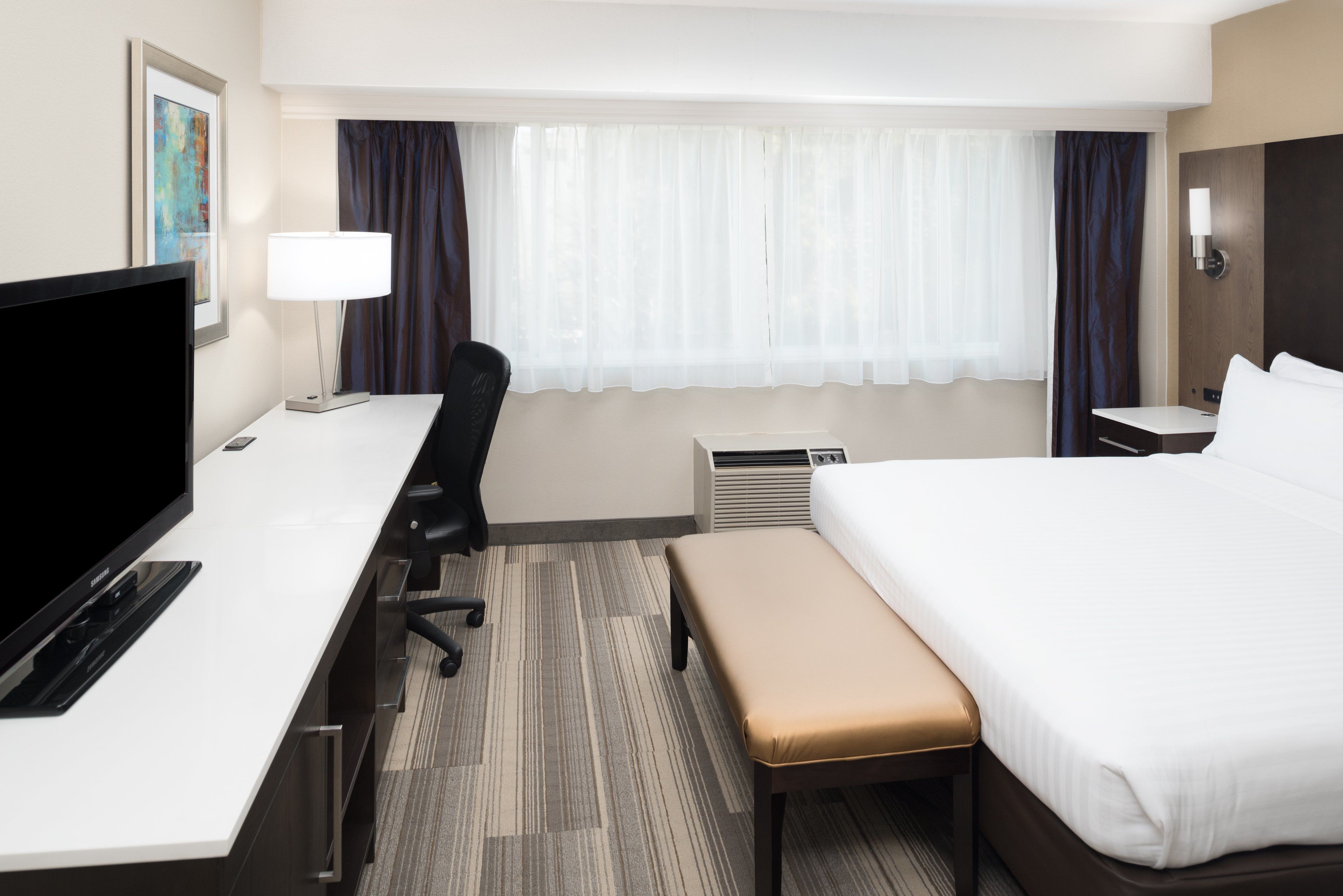 A hotel room with a bed, a desk with a TV, a chair, a bench, a lamp, and a window with curtains.
