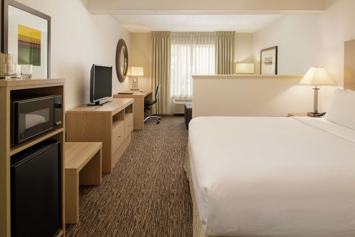 A hotel room with a bed, a desk with a computer, a television, a minibar, and a window with curtains.