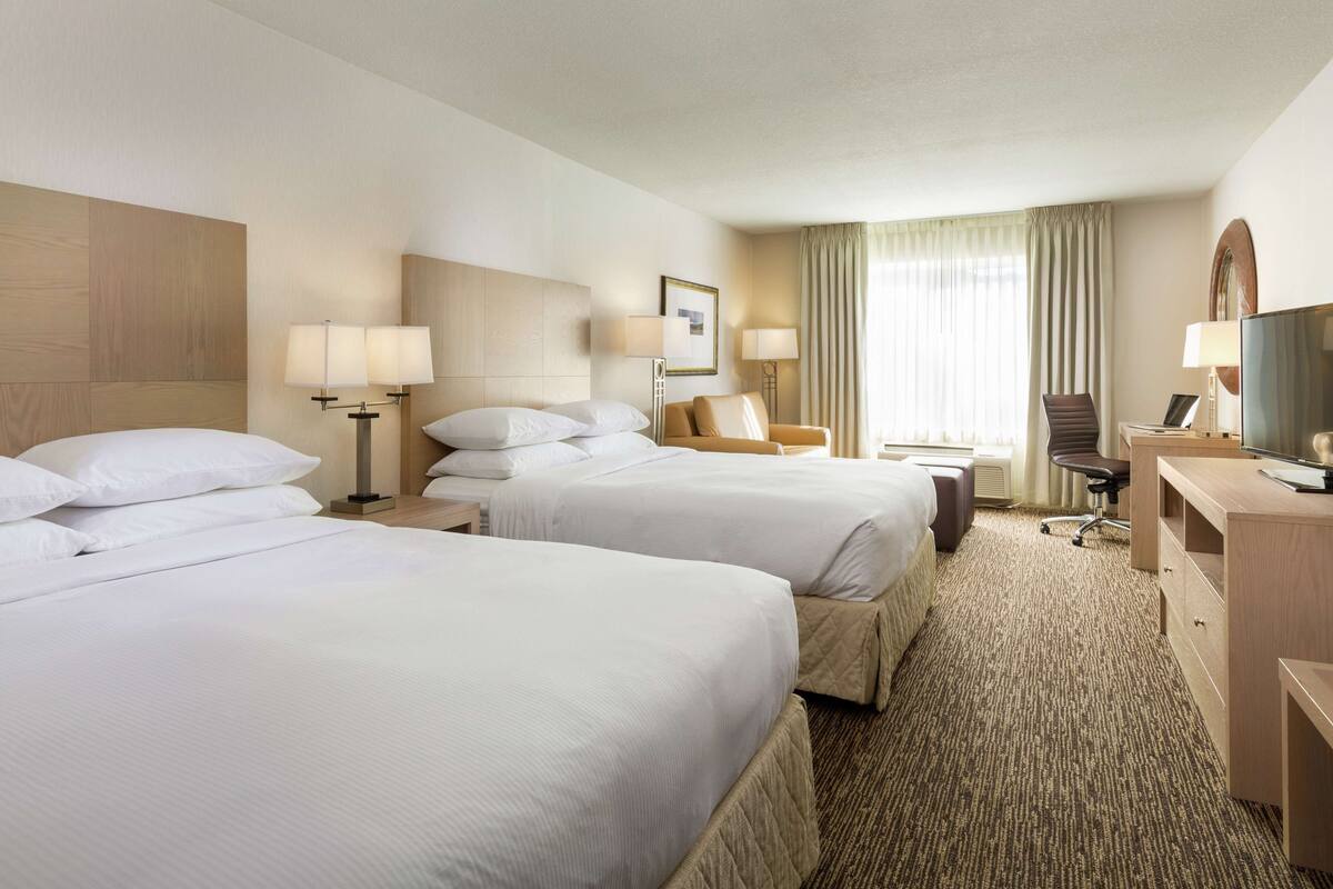 A hotel room with two beds, a desk, a chair, a TV, and a large window with curtains.