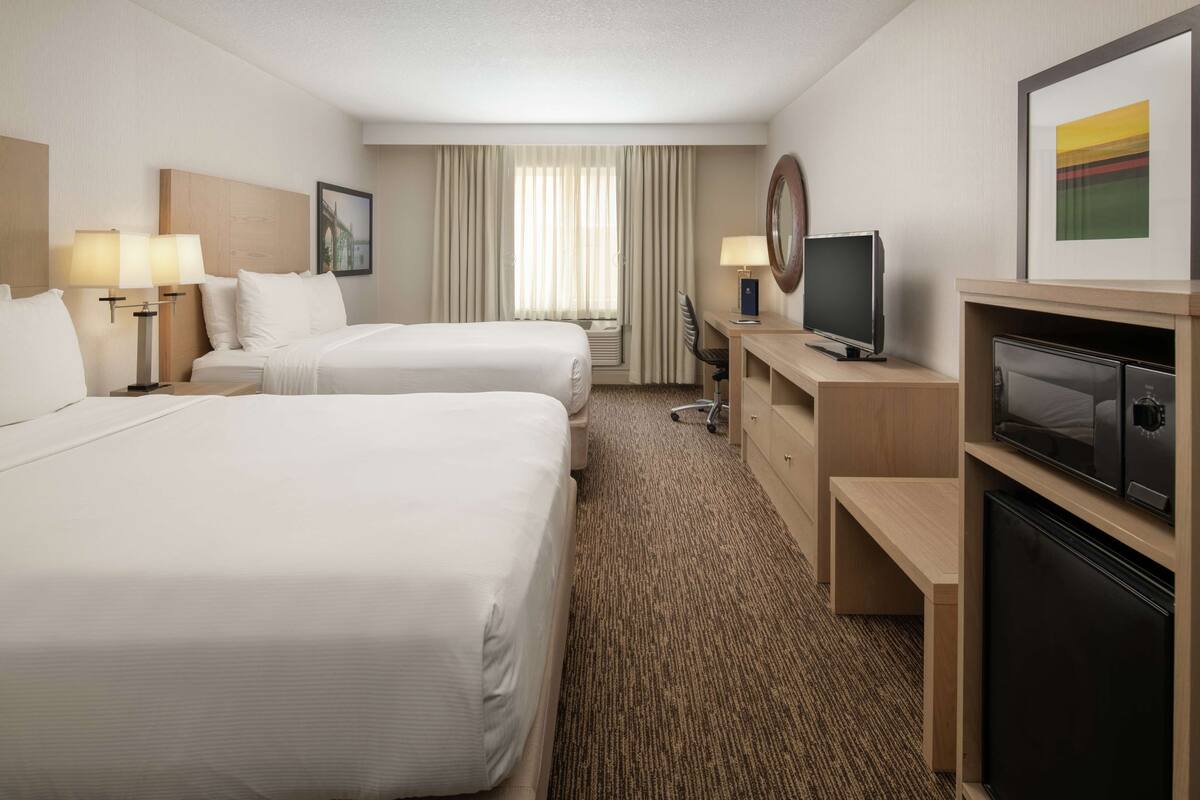 A hotel room with two beds, a desk with a television, a desk lamp, a chair, and a large window with curtains.