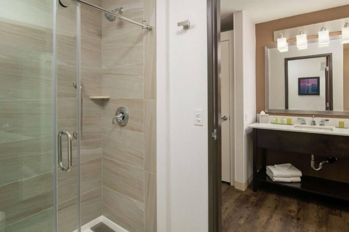 A modern bathroom with a glass shower enclosure, a dark vanity with a mirror, and a wooden floor.