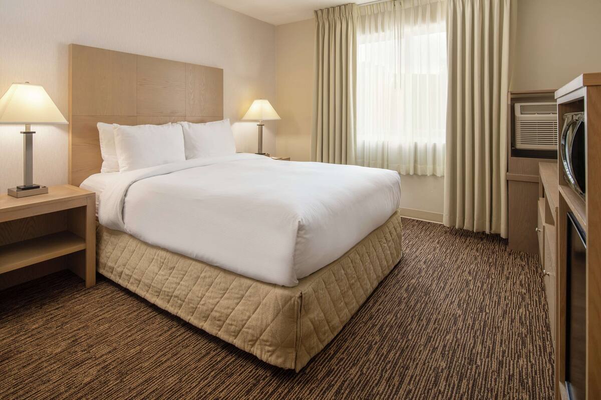 A hotel room with a large bed, two bedside lamps, a wooden headboard, a window with curtains, and a built-in TV.