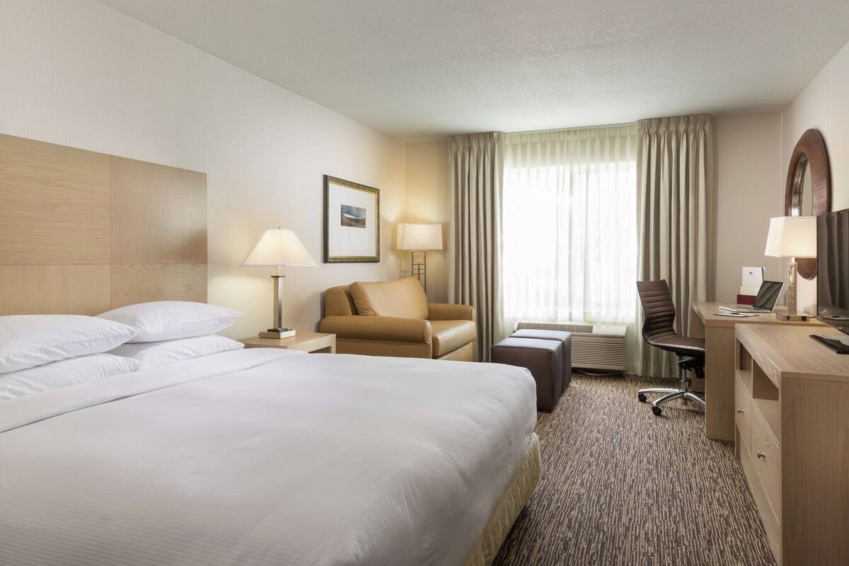 A hotel room with a large bed, a desk with a computer, a chair, a small table, and a window with curtains.
