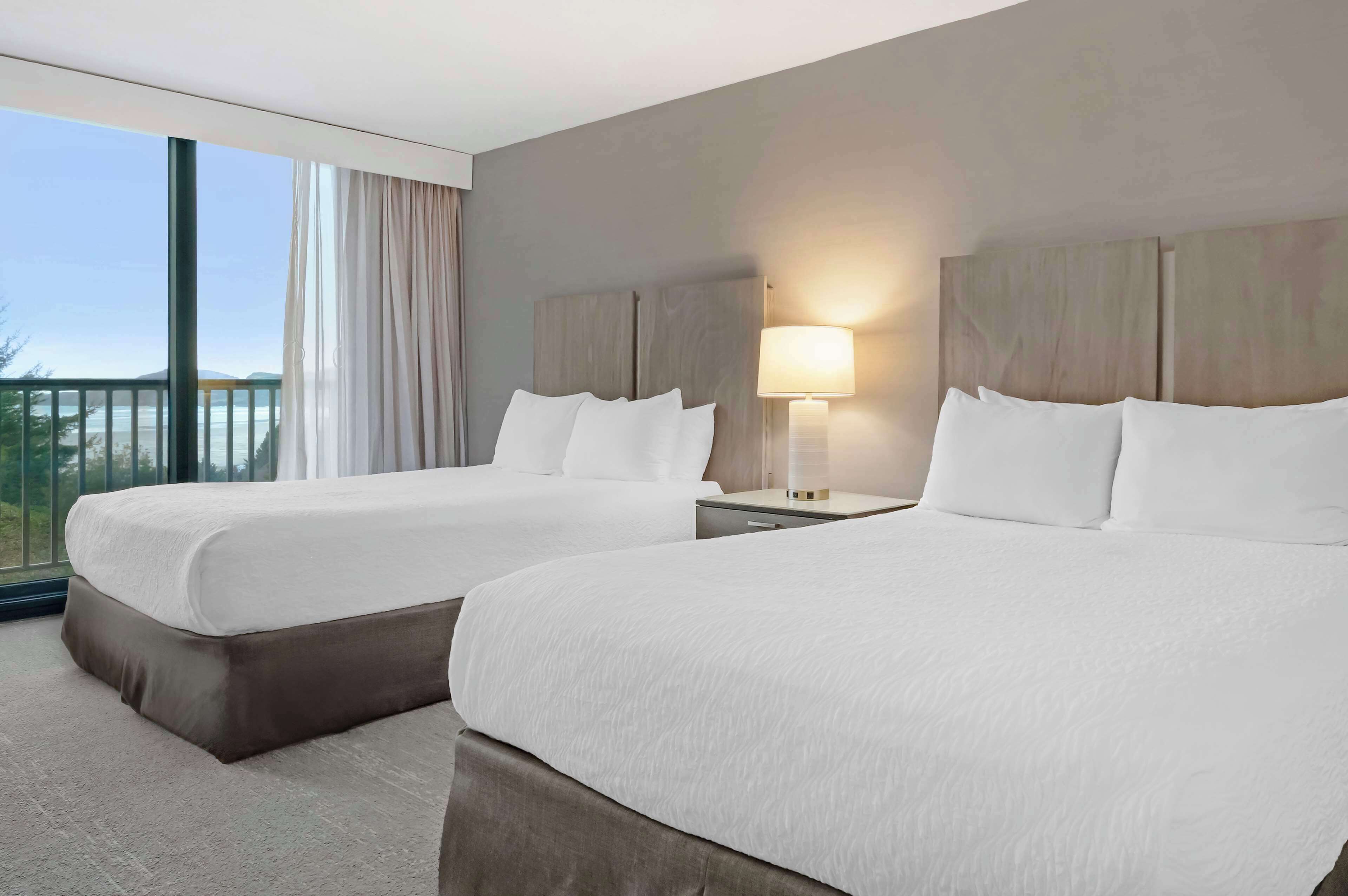 A hotel room with two beds, a large window with curtains, a nightstand with a lamp, and a view of the outdoors.