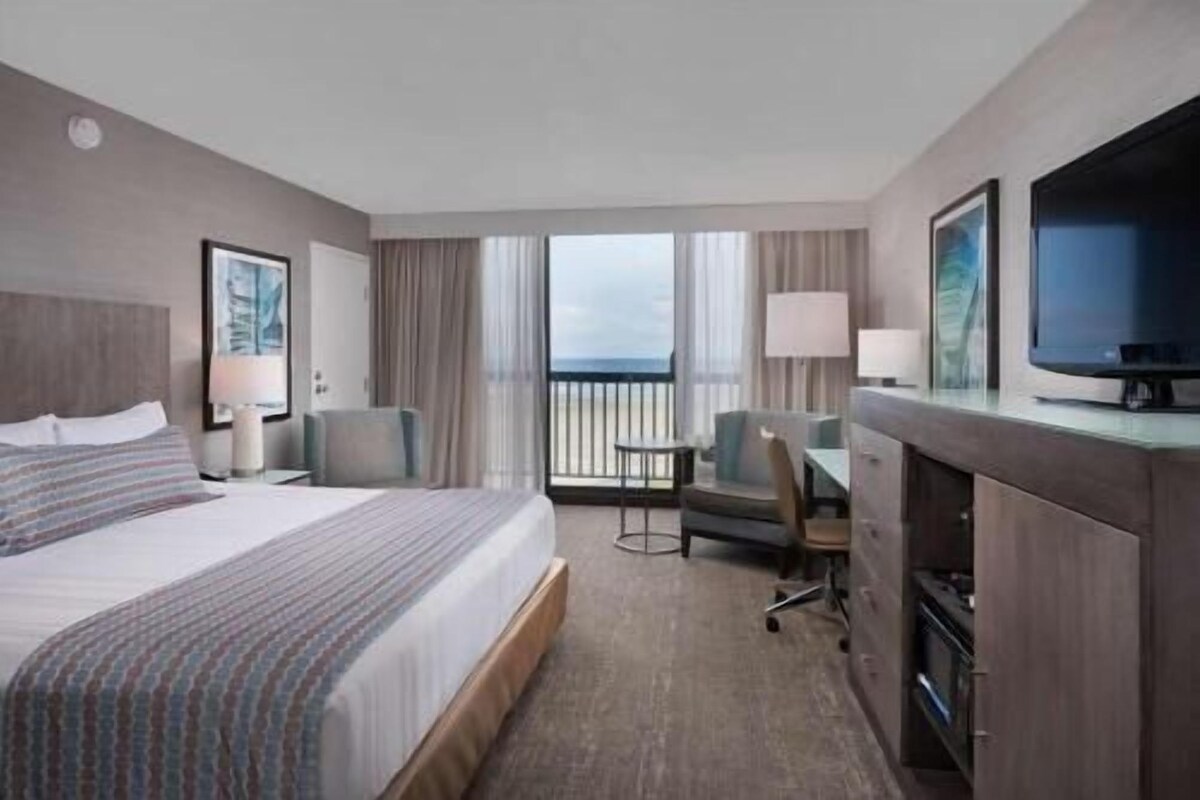 A hotel room with a bed, a desk, a chair, a TV, and a balcony with a view.