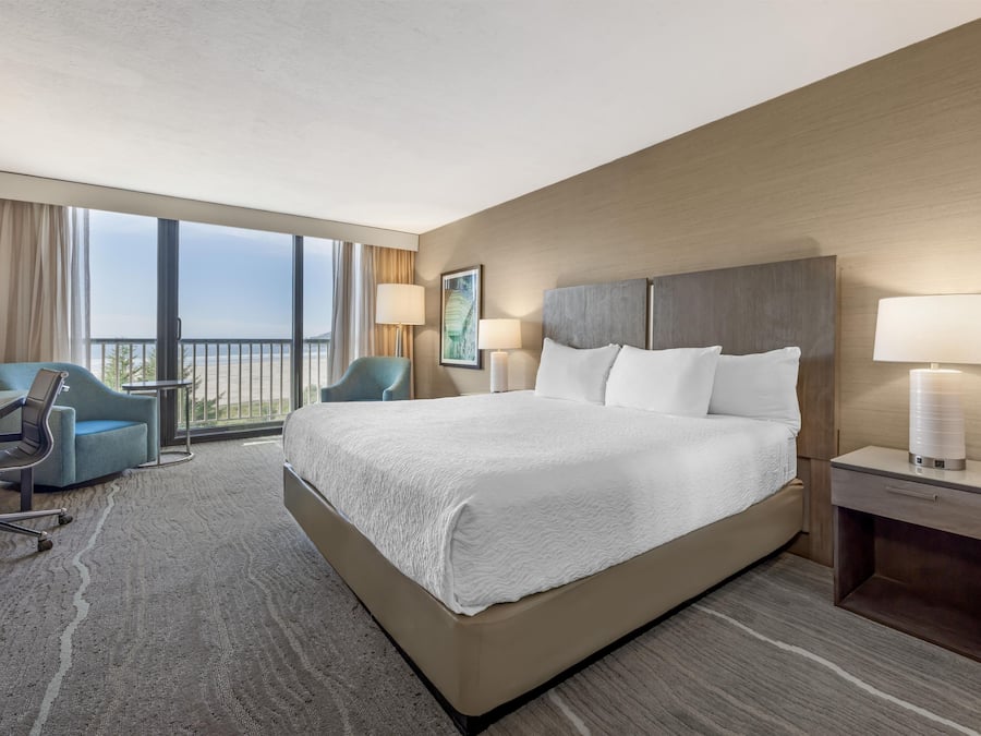 A hotel room with a large bed, a desk, a chair, a lamp, and a view of the outdoors.