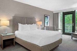 Habitación de hotel con cama, lámparas de noche, mesita de noche, una silla, una mesa y ventana con vistas al verde.
