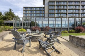 A patio area with Adirondack chairs and a fire pit, surrounded by a stone wall and a building with large windows in the background.
