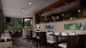 A restaurant with multiple tables and chairs, a bar area with glasses and bottles, and large windows offering a view of greenery.