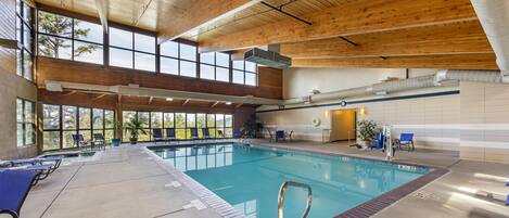 An indoor swimming pool with a wooden ceiling, clear windows, and lounge chairs.