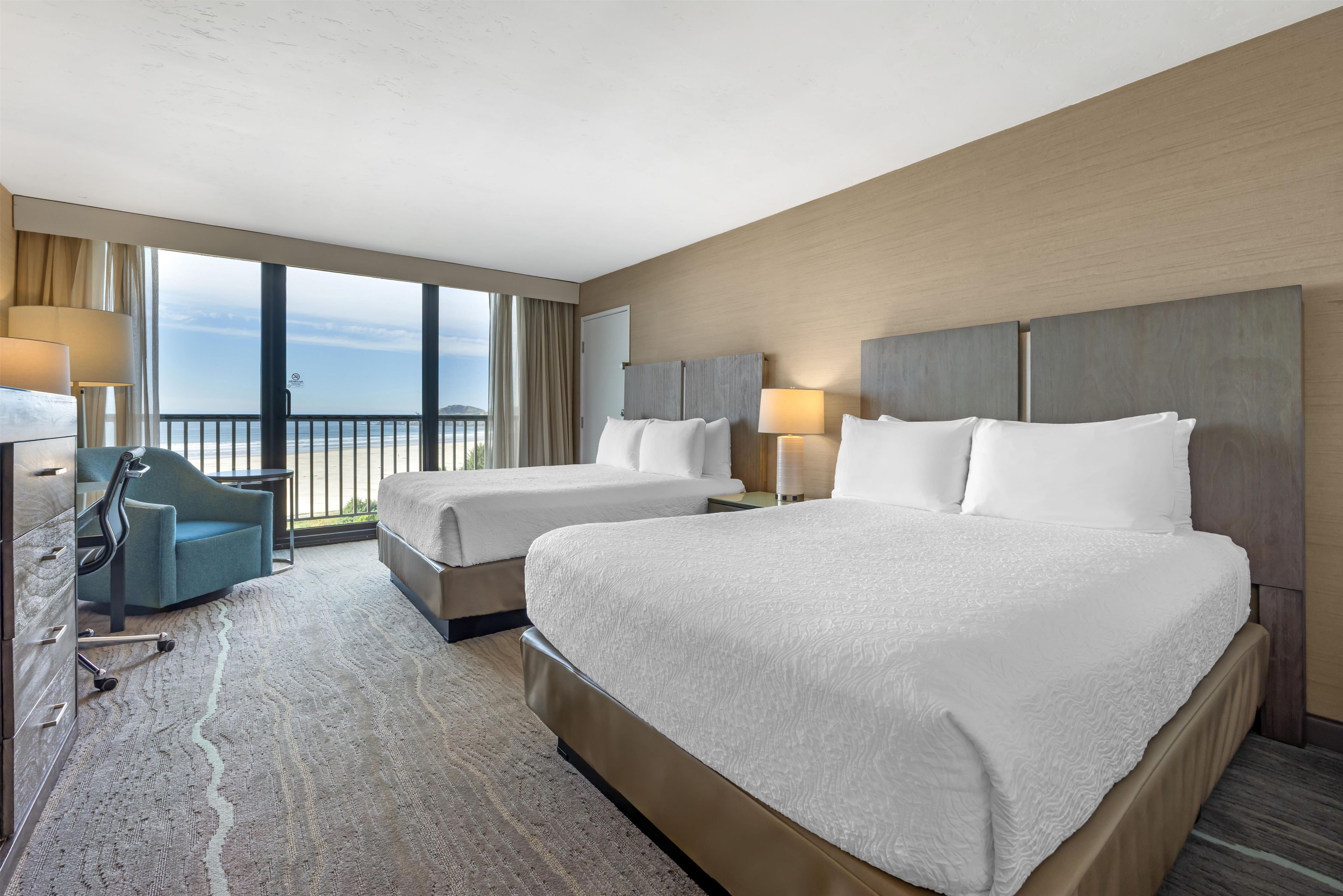 A hotel room with two beds, a desk, a chair, and a view of the ocean.