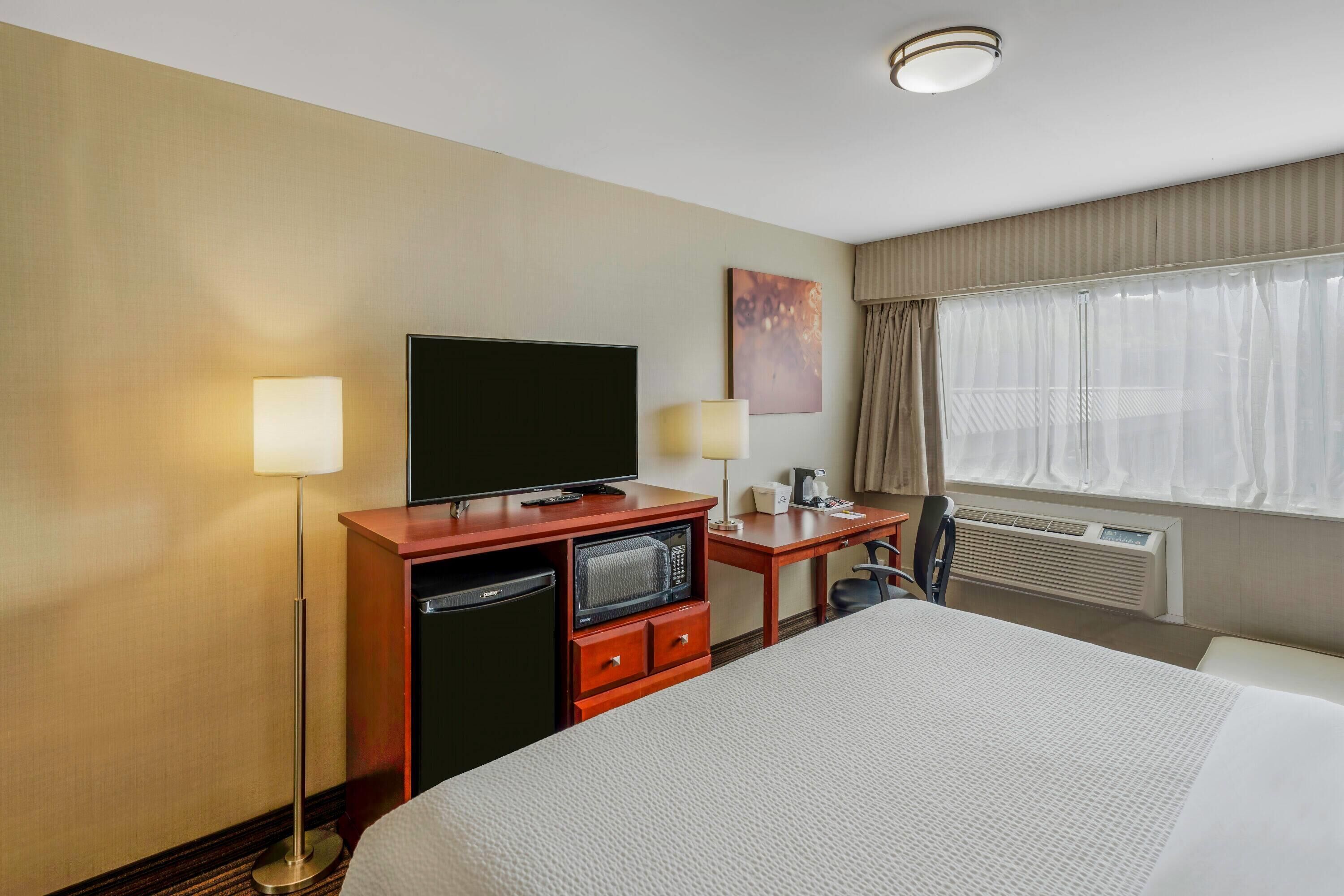 Une chambre d’hôtel avec un grand lit, un bureau avec une télévision, une lampe de bureau, un petit réfrigérateur et une fenêtre avec des rideaux.