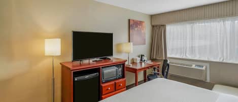 A hotel room with a large bed, a desk with a television, a desk lamp, a small refrigerator, and a window with curtains.