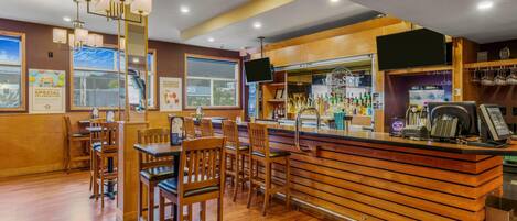 Uma área de bar com banquetas de madeira, um balcão com um caixa registradora e uma televisão montada acima do bar.