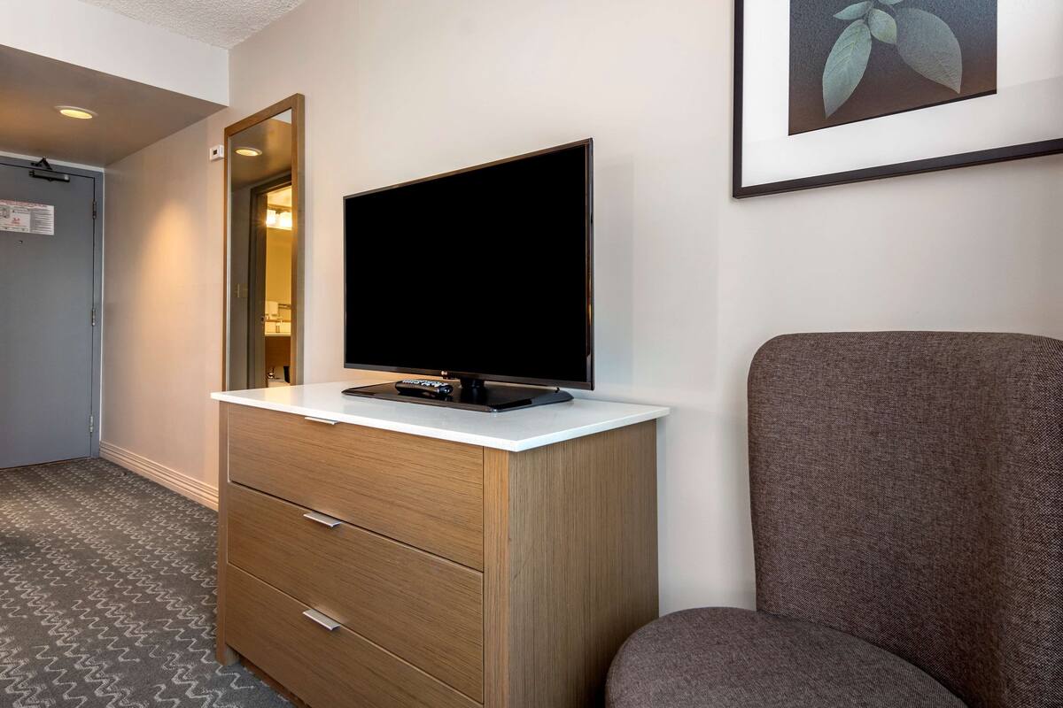 A hotel room with a flat-screen TV on a wooden cabinet, a gray armchair, and a framed picture on the wall.