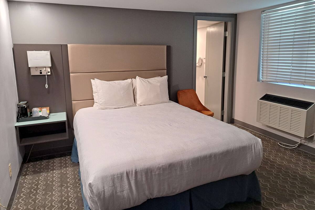 A hotel room with a large bed, a bedside table with a lamp, a chair, a window with blinds, and an air conditioning unit.