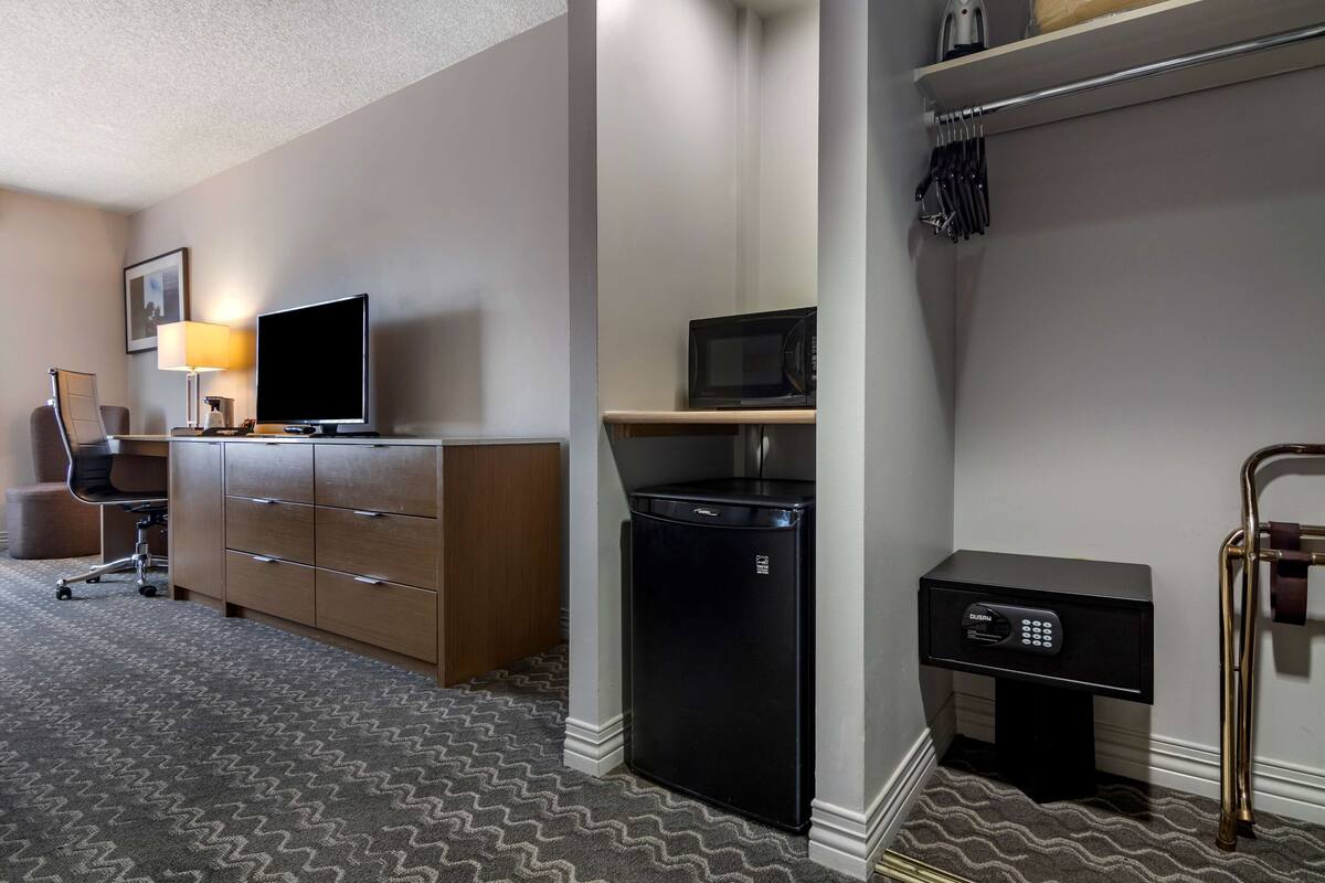 A hotel room with a wooden desk, a television, a microwave, a refrigerator, and a bedside table with a lamp.
