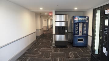 A hallway with a herringbone-patterned floor, a stainless steel refrigerator, a Pepsi vending machine, and a glass elevator.