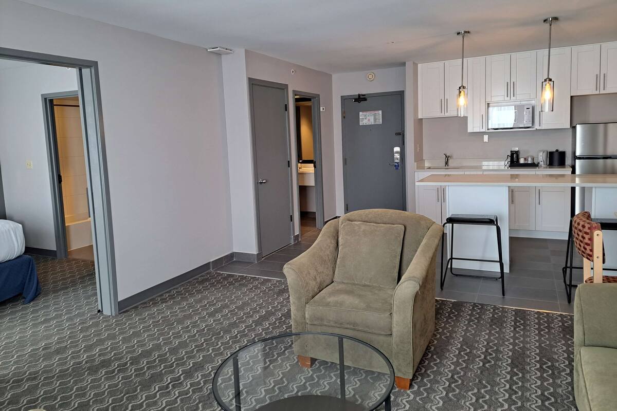 A modern apartment with a kitchenette, a beige armchair, a glass coffee table, and a patterned carpet.