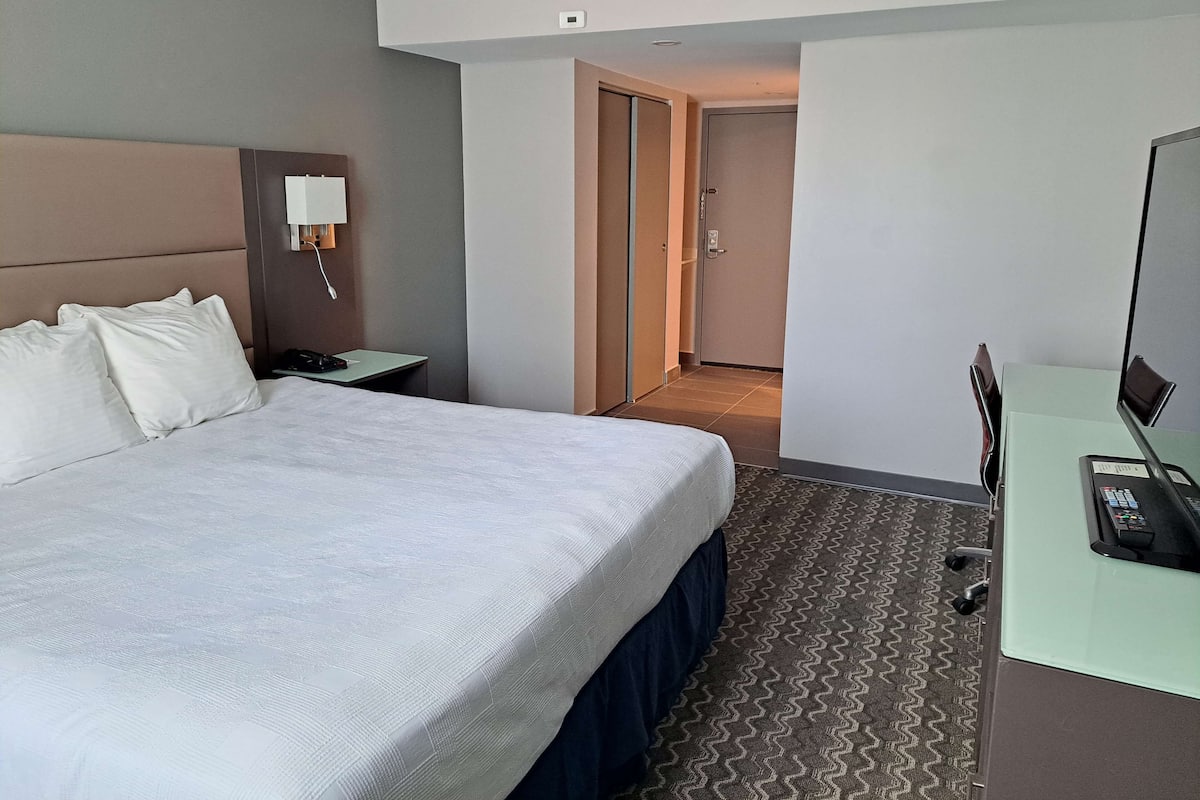 A hotel room with a large bed, a desk with a computer, a nightstand, and a wall-mounted lamp.