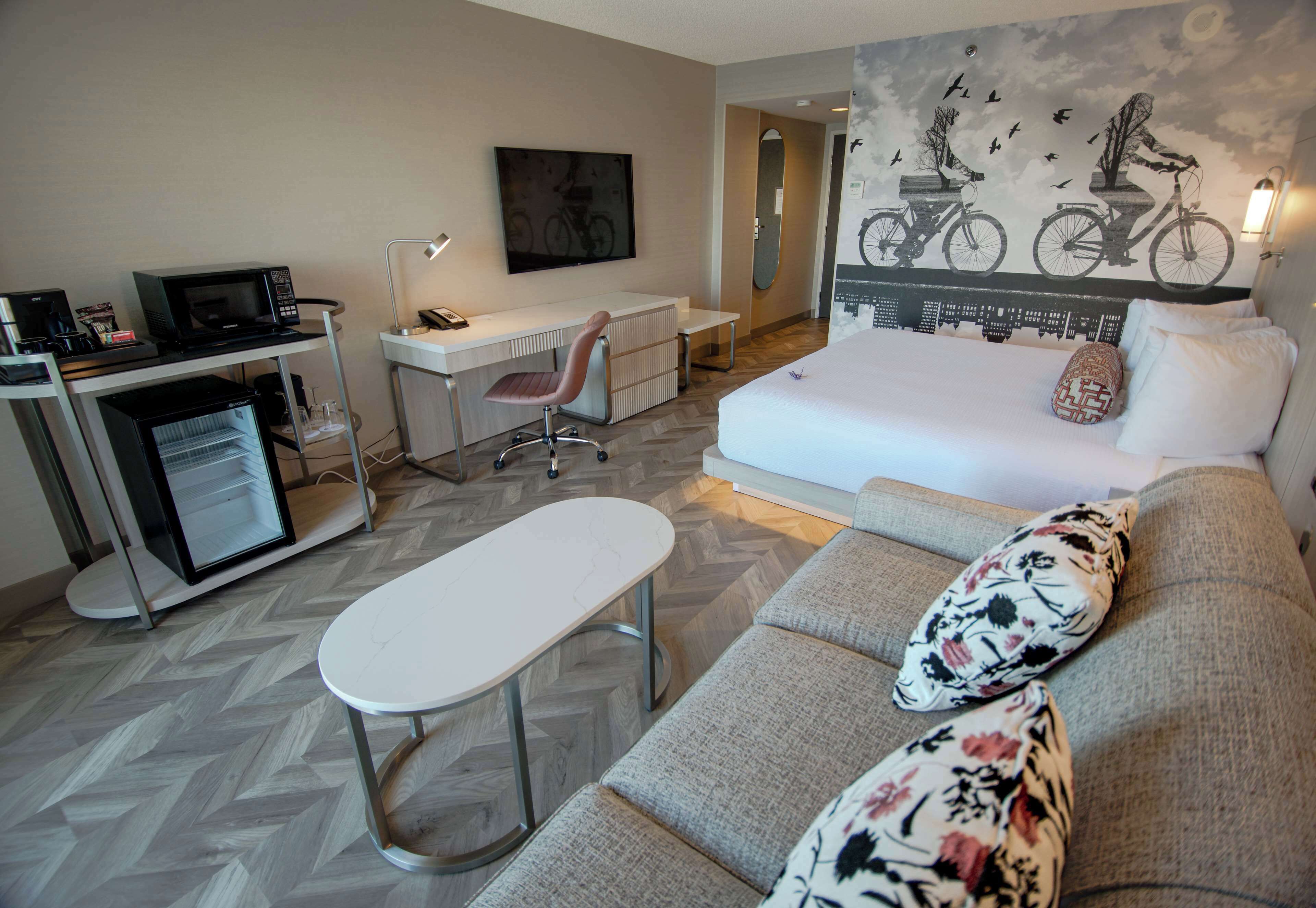 A modern hotel room with a sofa, a small desk, a chair, a flat-screen TV, and a wall-mounted mirror.