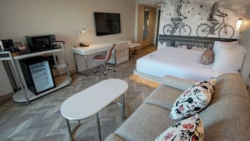A modern hotel room with a sofa, a small desk, a chair, a flat-screen TV, and a wall-mounted mirror.