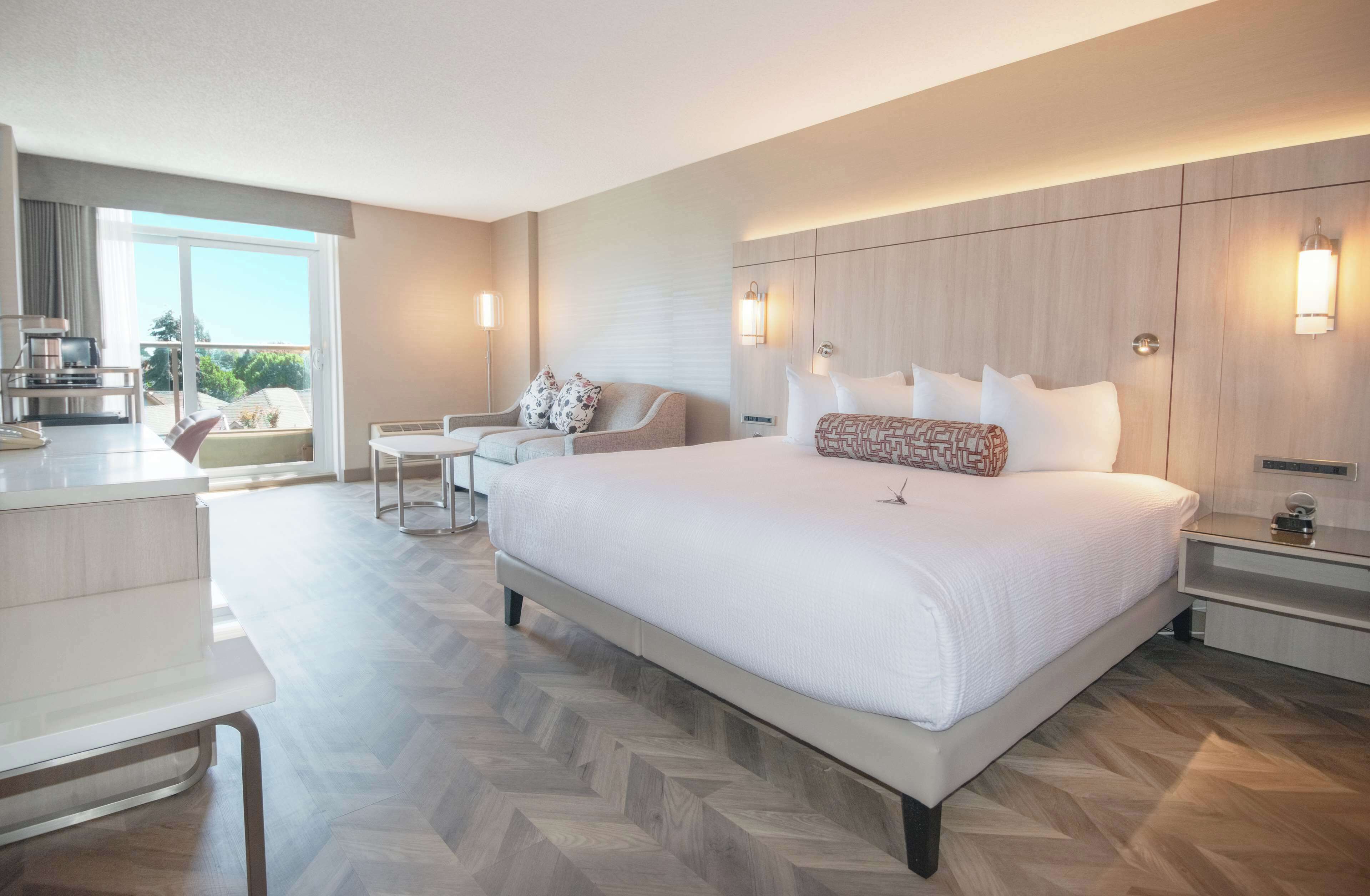 Ein modernes Hotelzimmer mit einem großen Bett, einem Schreibtisch, einem Sessel und Blick ins Freie.