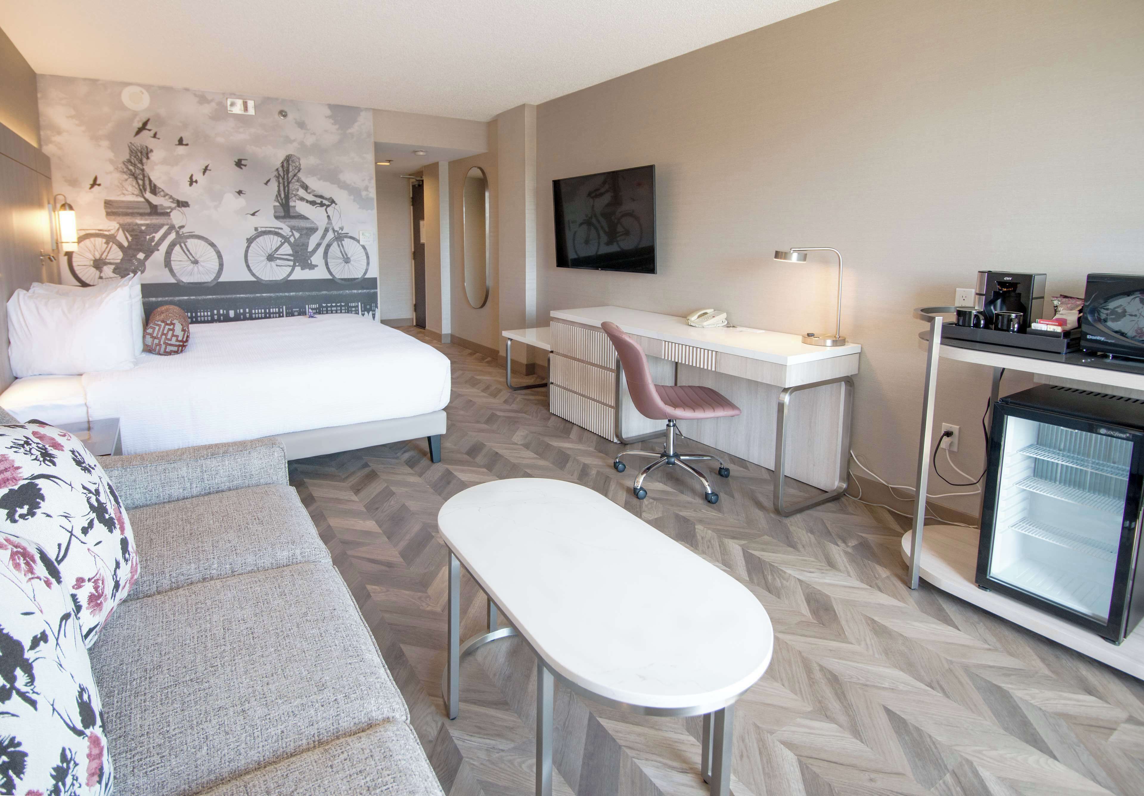 A hotel room with a bed, a desk, a chair, a small table, a refrigerator, and a wall with a mural.
