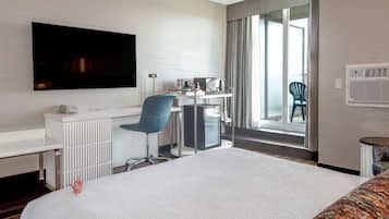 Habitación de hotel con una cama grande, un escritorio con computadora y un televisor de pantalla plana, una silla y vistas al exterior a través de un ventanal amplio.