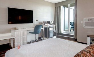 Habitación de hotel con una cama grande, un escritorio con computadora y un televisor de pantalla plana, una silla y vistas al exterior a través de un ventanal amplio.