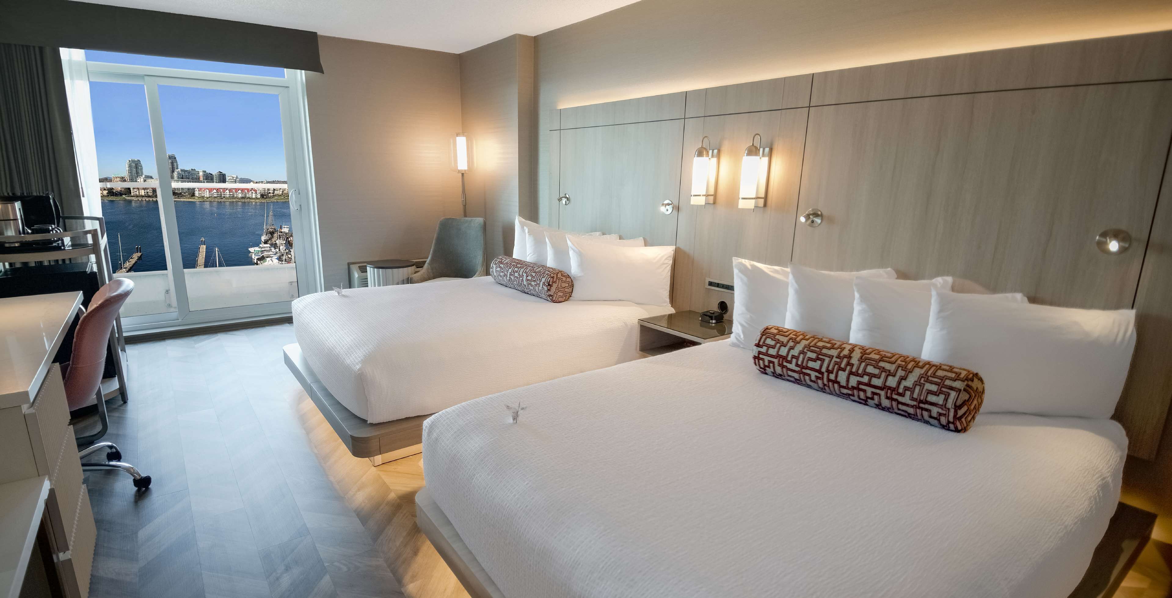 Habitación de hotel con dos camas, un escritorio, una silla y vistas a un puerto deportivo y al horizonte urbano.