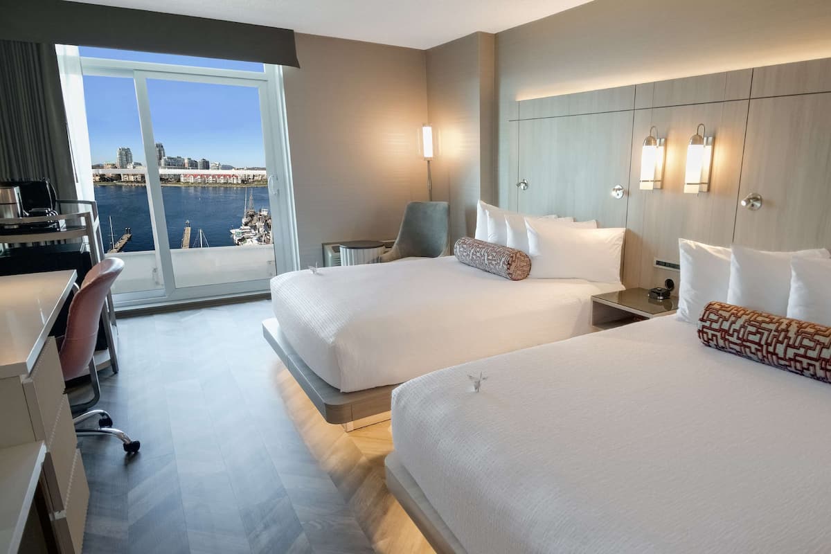 Hotellihuone, jossa on kaksi sänkyä, työpöytä, tuoli ja näkymä venesatamaan ja kaupungin siluettiin.