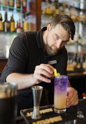 Un cantinero preparando una bebida en la barra, con una estantería repleta de botellas al fondo.