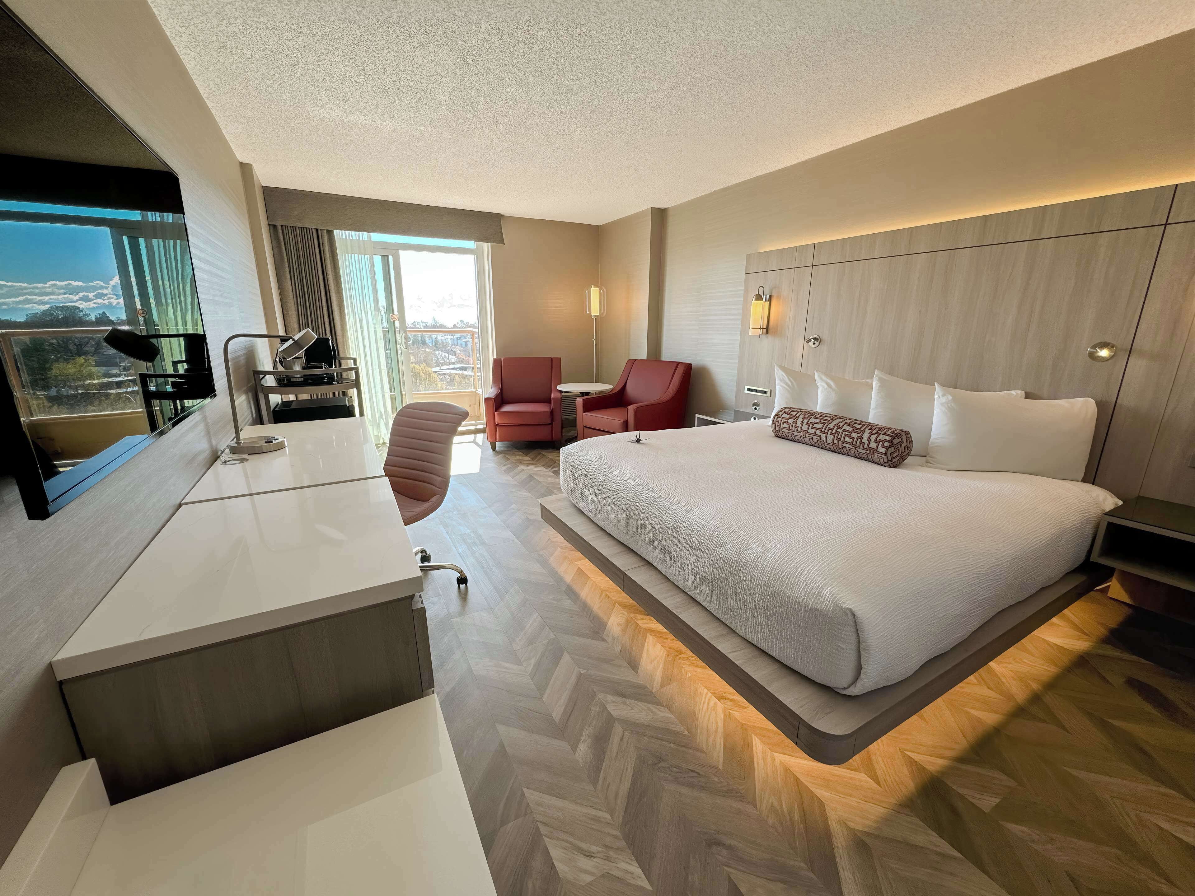 Una habitación de hotel moderna con una cama grande, un escritorio con una silla, un sillón rojo y vistas de la ciudad a través de la ventana.