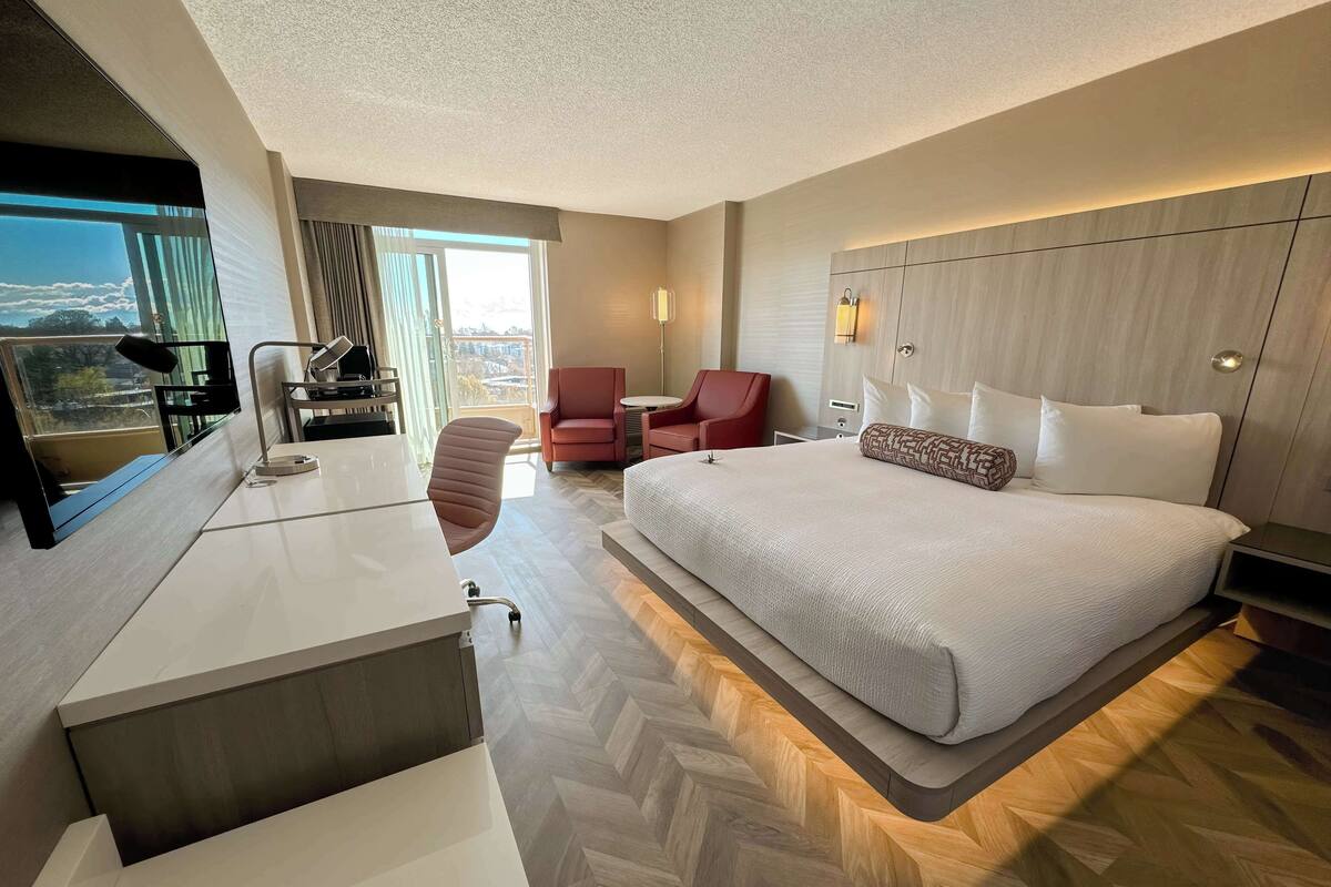 Moderni hotellihuone, jossa on suuri sänky, työpöytä tuolilla, punainen nojatuoli ja ikkunasta avautuva näkymä kaupunkiin.