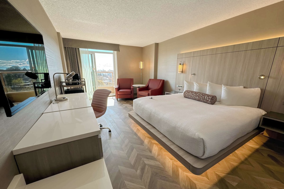 Una habitación de hotel moderna con una cama grande, un escritorio con una silla, un sillón rojo y vistas de la ciudad a través de la ventana.