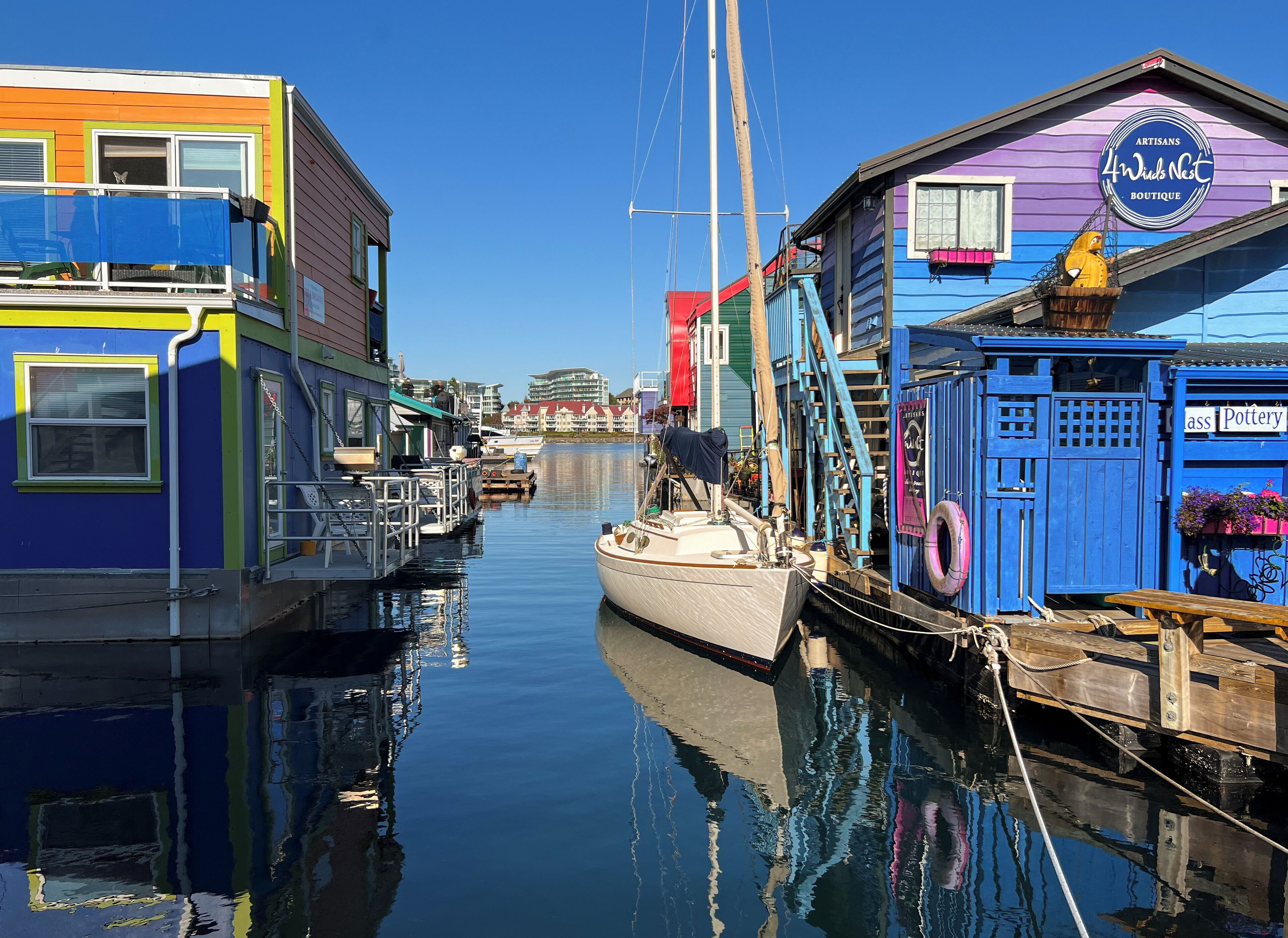 A narrow waterway with colorful buildings on either side, including a blue building with a sign for 'Artisans 4 Winds Nest Boutique' and a 'Pottery' sign.