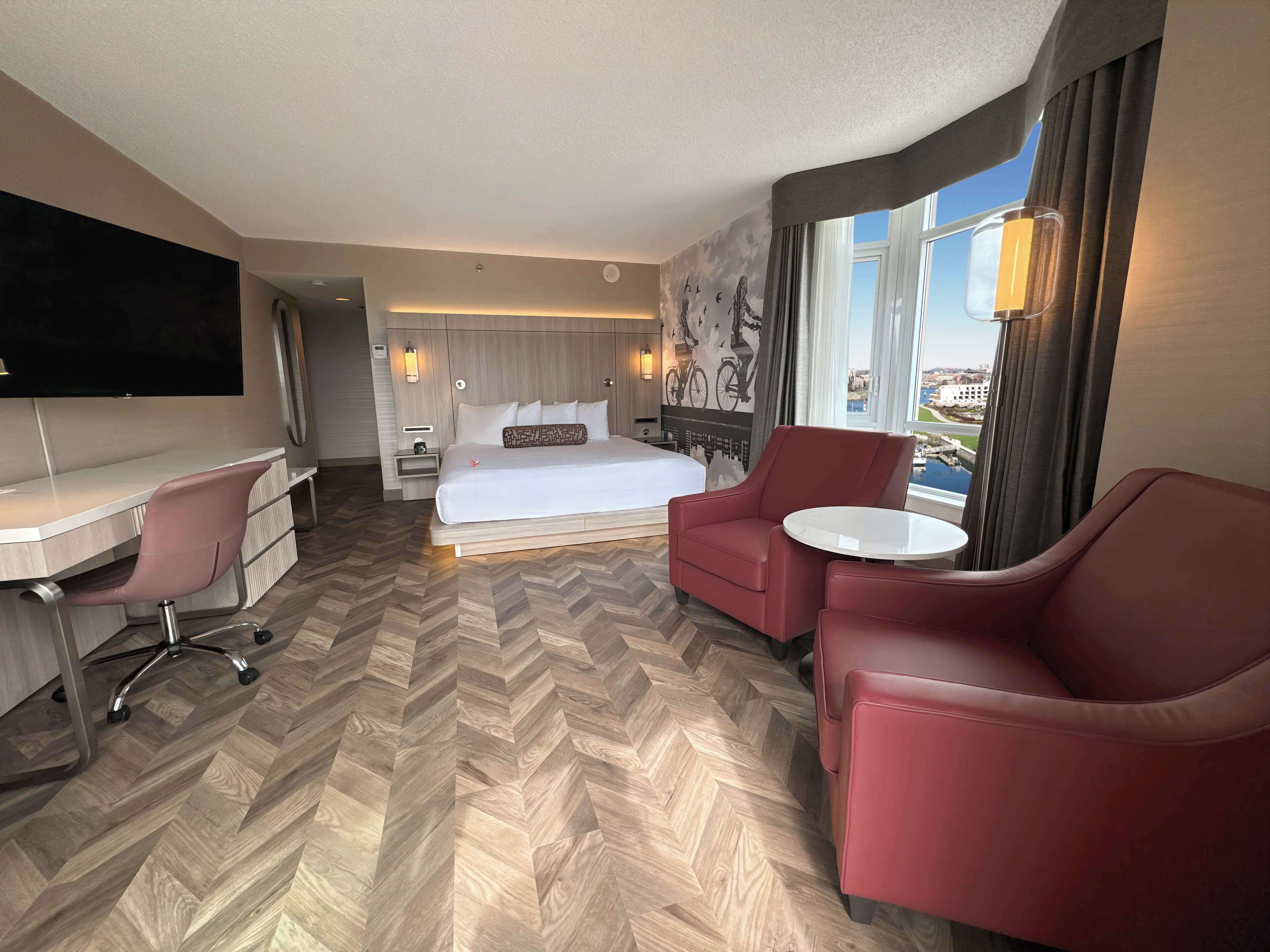 Habitación de hotel con cama, escritorio, dos sillas, televisor y ventana grande con vista a la ciudad.