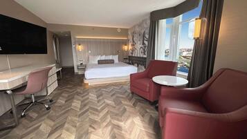 Habitación de hotel con cama, escritorio, dos sillas, televisor y ventana grande con vista a la ciudad.