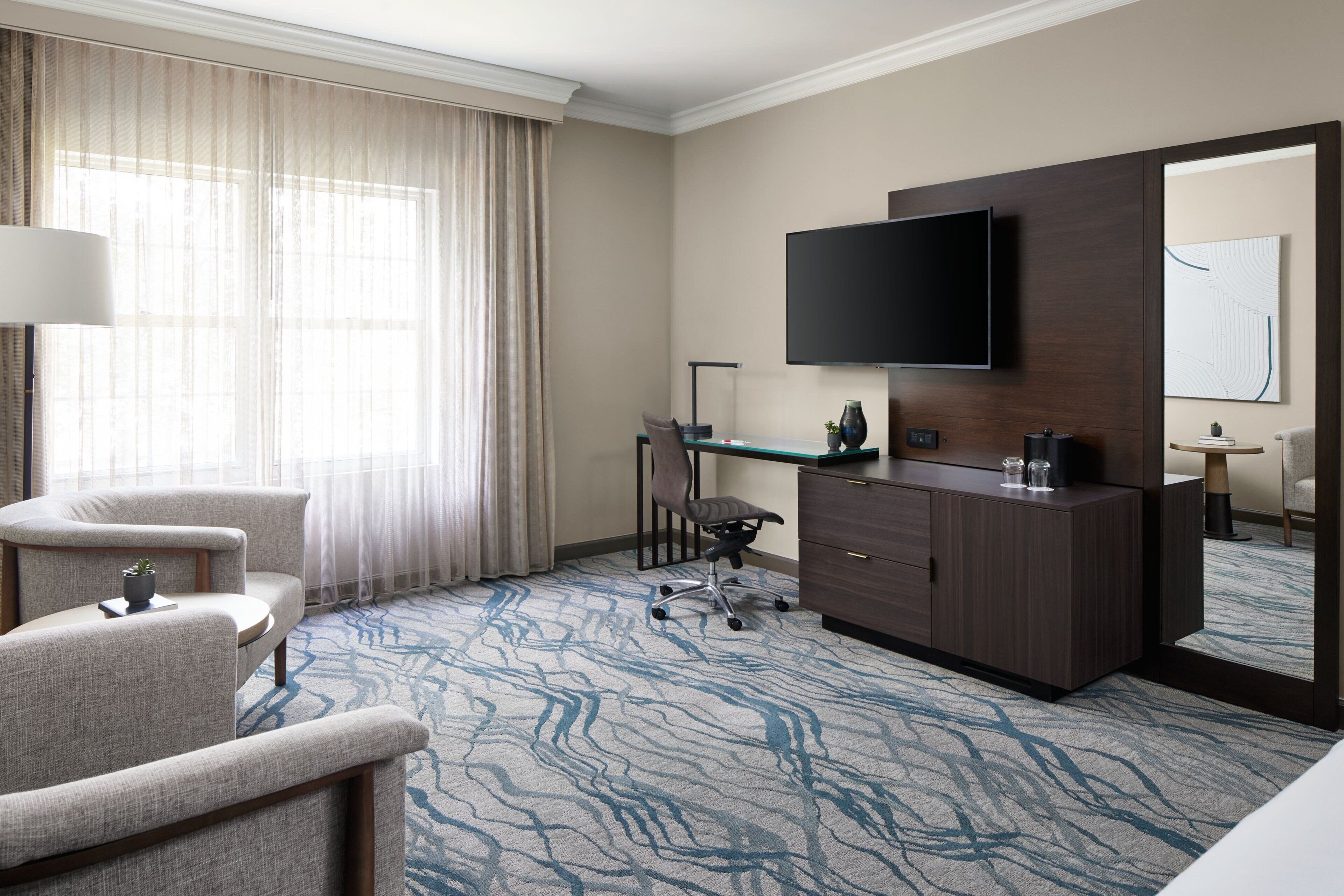 Une chambre d’hôtel dotée d’un grand téléviseur à écran plat fixé au mur, d’un bureau avec une chaise, d’un canapé et d’une moquette à motifs.