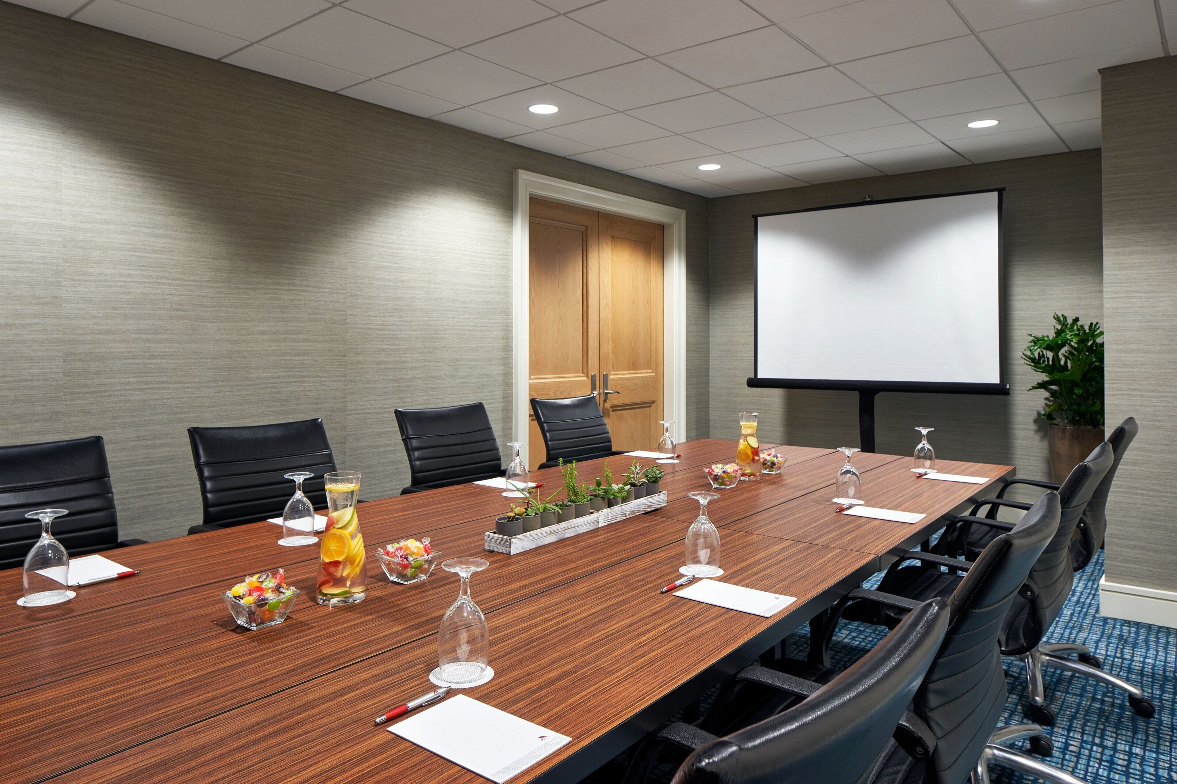 A conference room with a long wooden table, black chairs, a projector screen, and a potted plant.