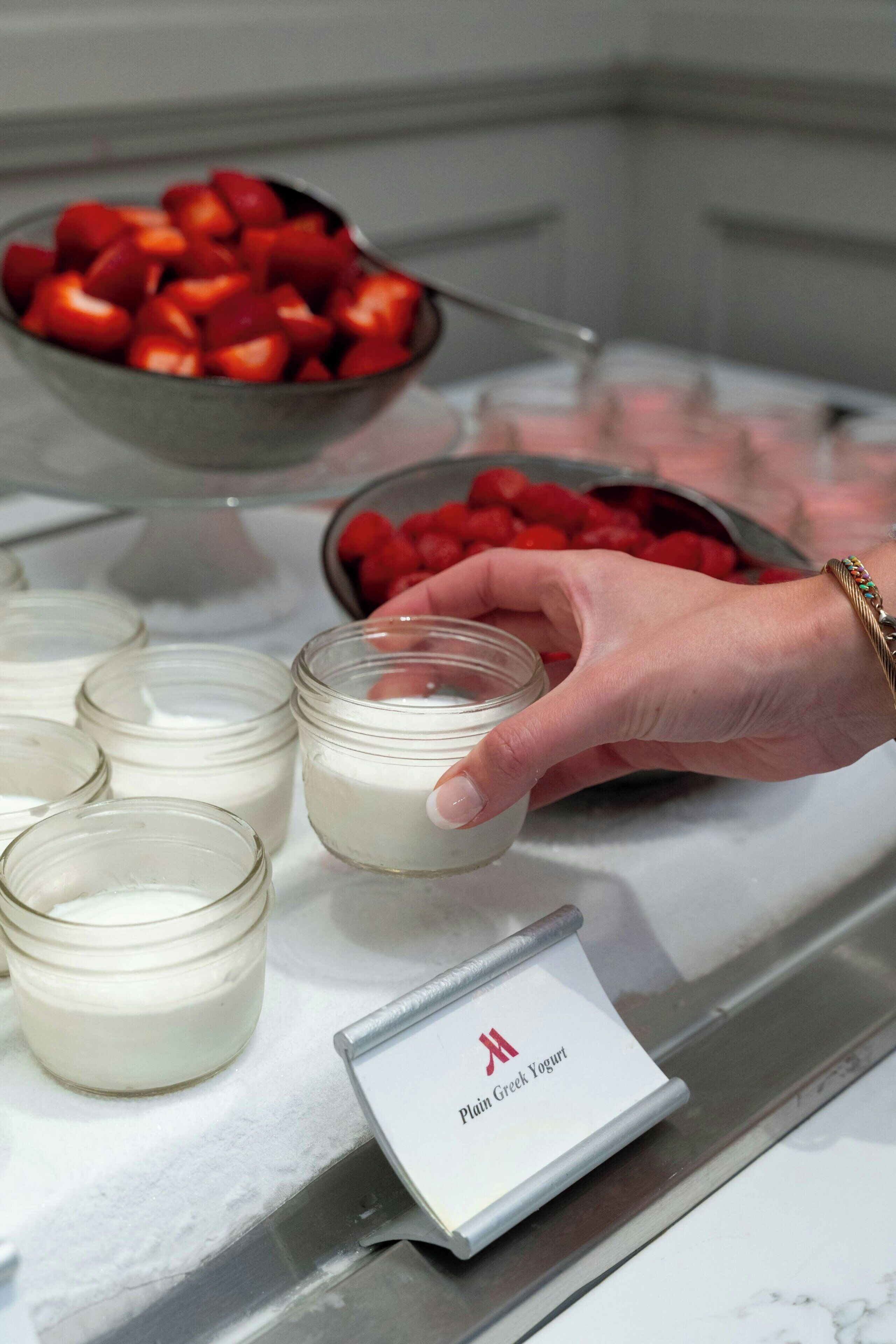 A hand reaching for a jar of yogurt with a label reading "Plain Greek Yogurt" on a counter.