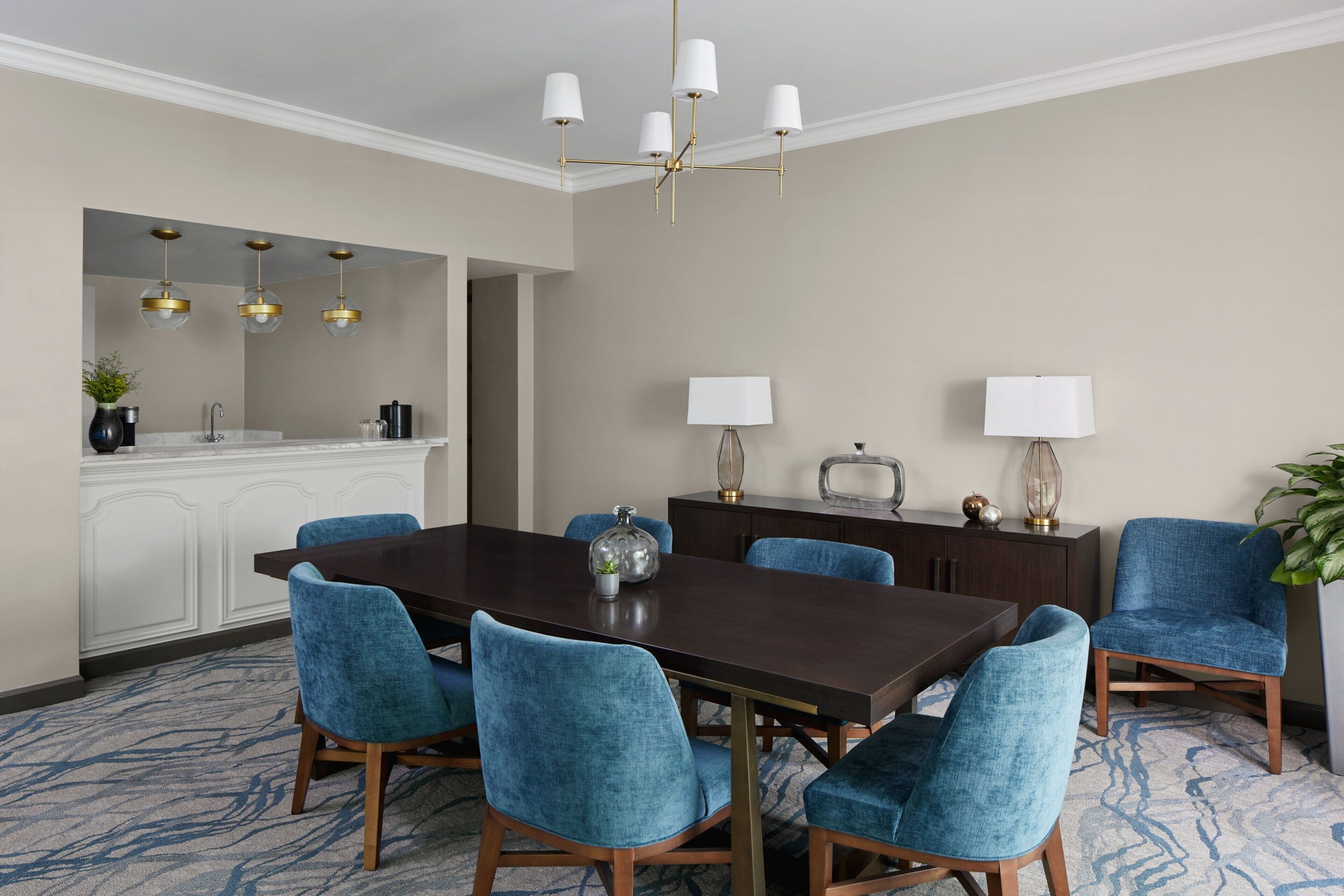 Un coin repas avec une table en bois foncé, six chaises bleues à revêtement, un lustre et un meuble blanc avec un évier.