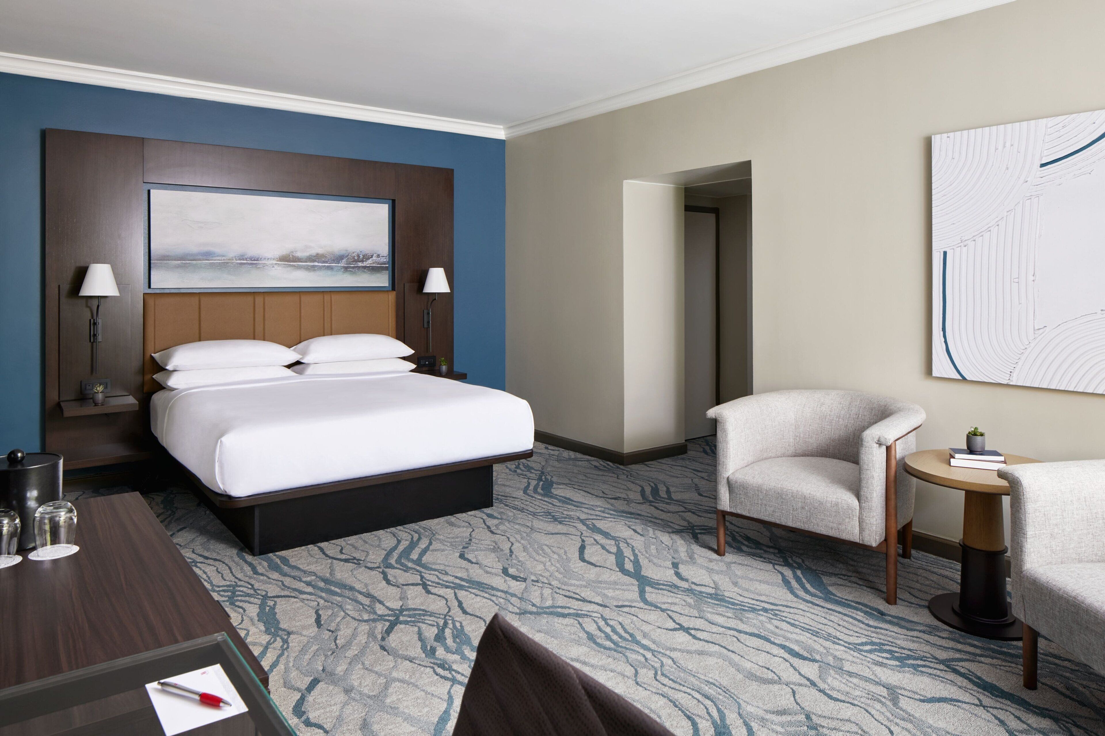A hotel room with a large bed, two bedside lamps, a small table with a lamp, a chair, and a wall-mounted artwork.