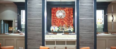 A bar area with a patterned wall, marble countertop, and orange chairs.