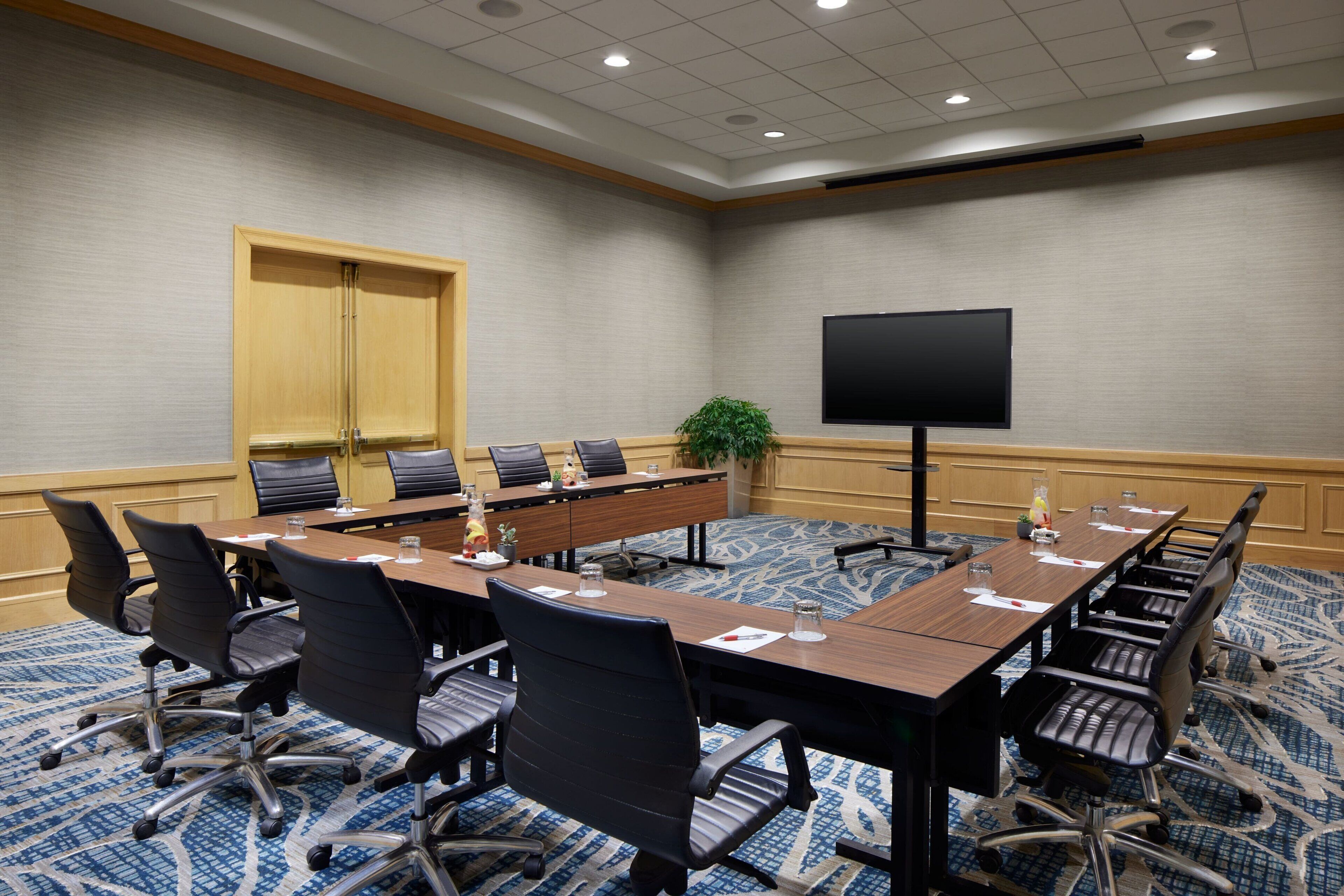A conference room with a long table, multiple chairs, a flat-screen TV, and a plant.