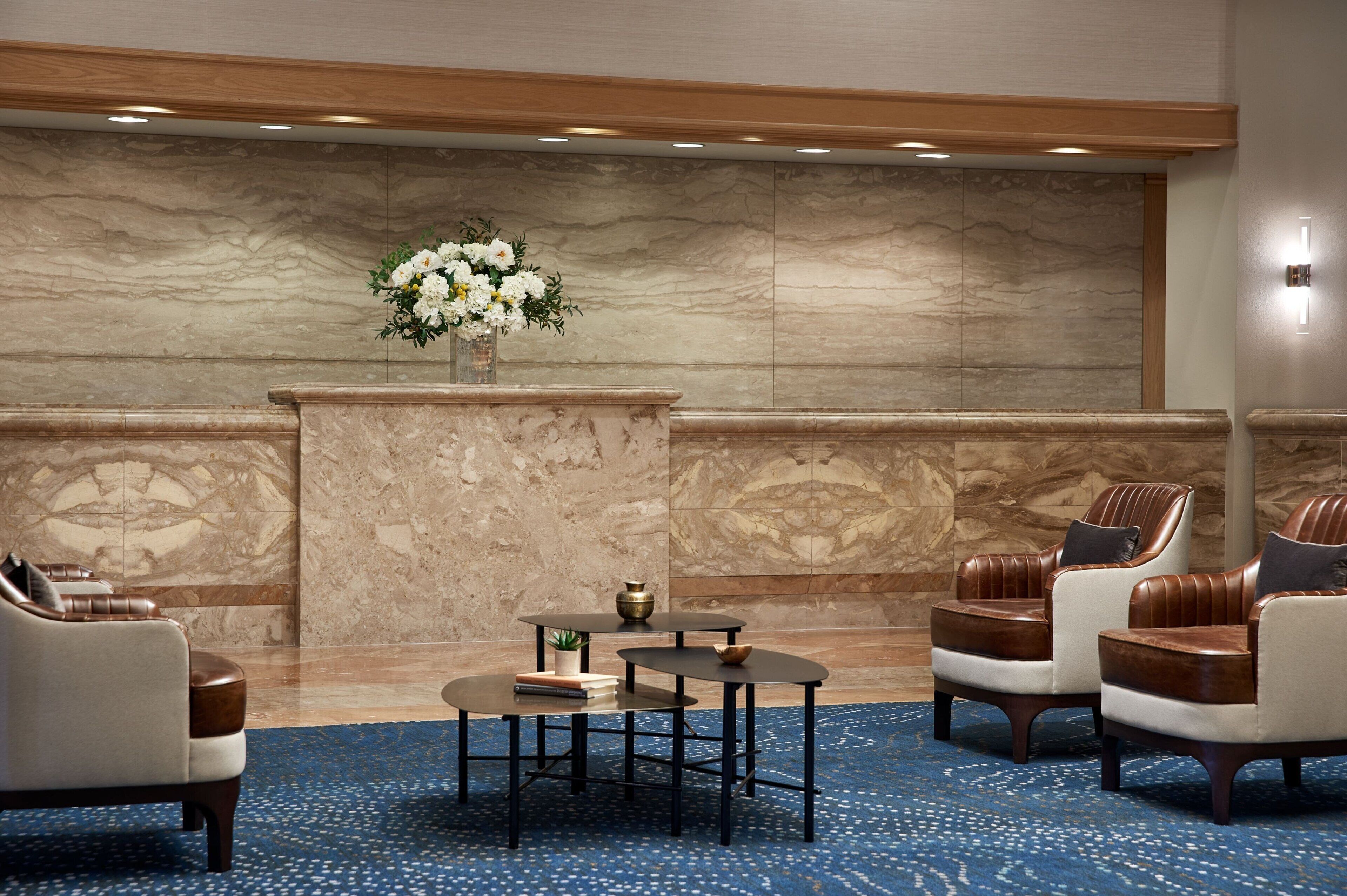 A lobby with a marble wall, a vase of flowers, leather chairs, a small table with books, and a blue carpet.