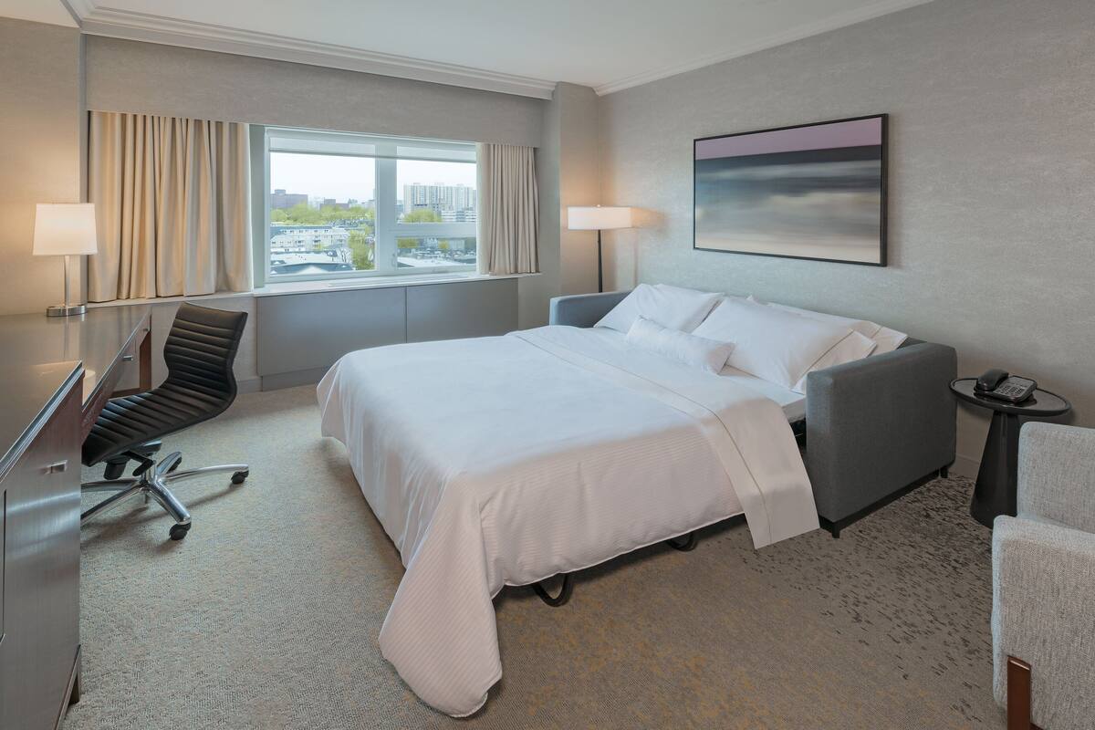 A hotel room with a large bed, a desk, a chair, and a view of the city through the window.